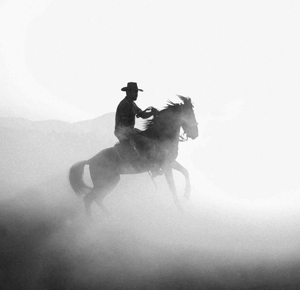 A dramatic black and white silhouette of a cowboy riding a horse in a dusty landscape.